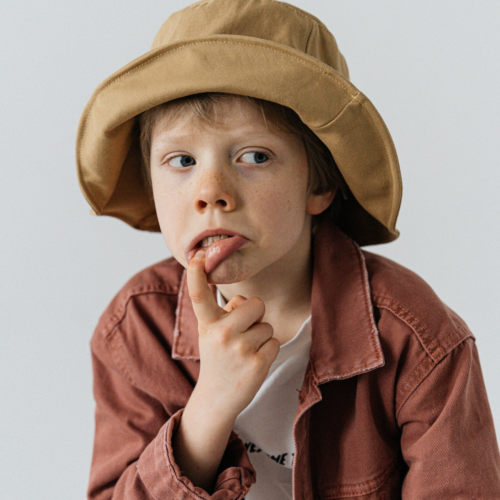 Niño pelirrojo con pecas y ojos azules, usando un sombrero de cubo, con el dedo en la boca mirando hacia la derecha, simbolizando la curiosidad